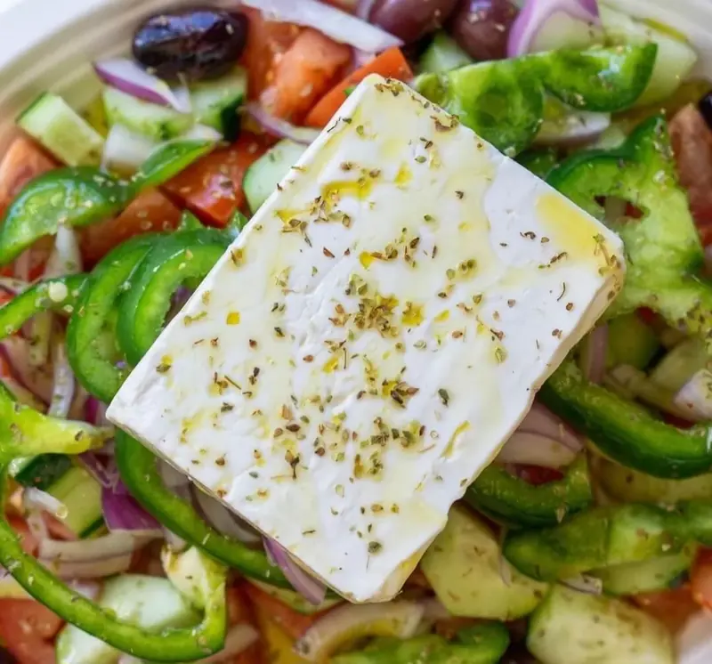 Greek salad under a slab of feta with green peppers and olives